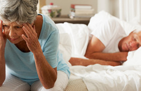 Worried Senior Woman Sits On Bed Whilst Husband Sleeps